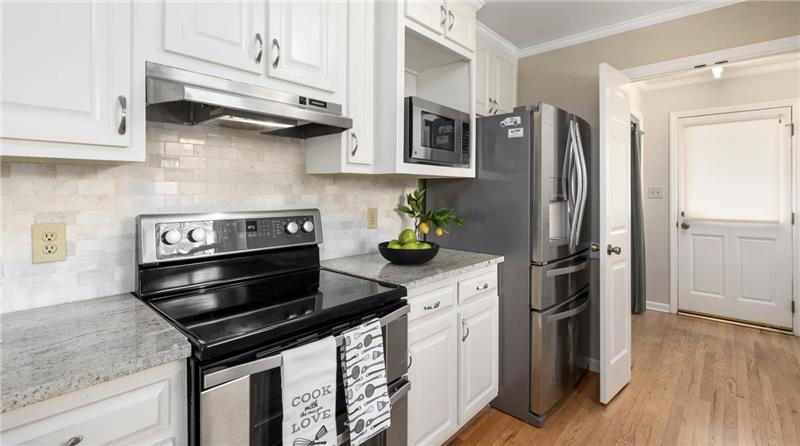 Modern kitchen featuring stainless steel appliances, including a stove and refrigerator, with white cabinetry and granite countertops, showcasing a bowl of fresh fruit and a dish towel with cooking-themed text.